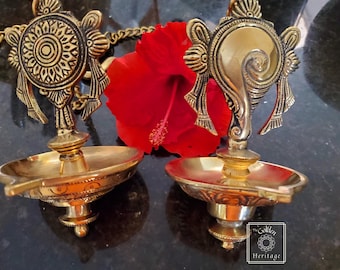 Puede incluir: Dos lámparas de aceite colgantes de color dorado con diseños intrincados. Las lámparas están suspendidas por una cadena y tienen un cuenco decorativo para aceite. Las lámparas están delante de una flor de hibisco roja.