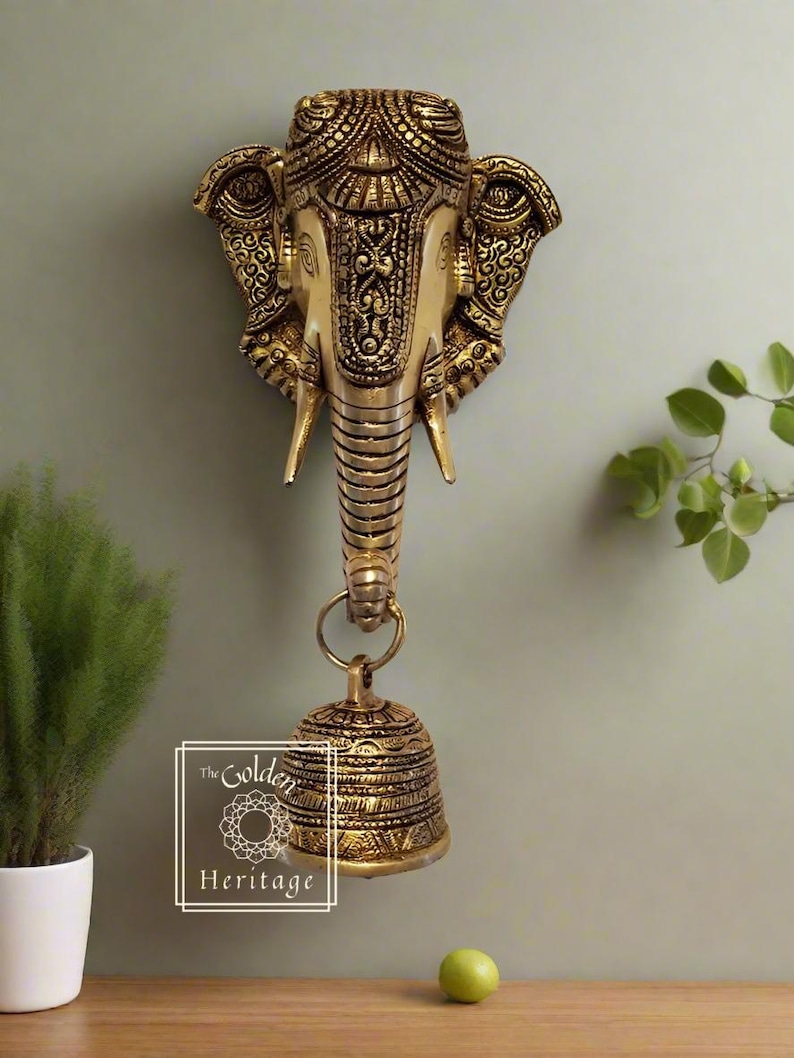 Puede incluir: Una decoraci&oacute;n de pared de metal dorado con una cabeza de elefante detallada con una campana colgando de su trompa. La cabeza de elefante tiene dise&ntilde;os intrincados y la campana tambi&eacute;n est&aacute; decorada con patrones. La decoraci&oacute;n de pared est&aacute; etiquetada como "The Golden Heritage".