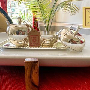 Puede incluir: Un plato de servir de plata con forma de manzana con tapa, sentado en una bandeja de plata con un borde de cuentas. El plato est&aacute; sobre una bandeja ovalada blanca, que est&aacute; sobre un mantel individual rojo. El texto "The Golden Heritage" es visible en la esquina superior derecha de la imagen.