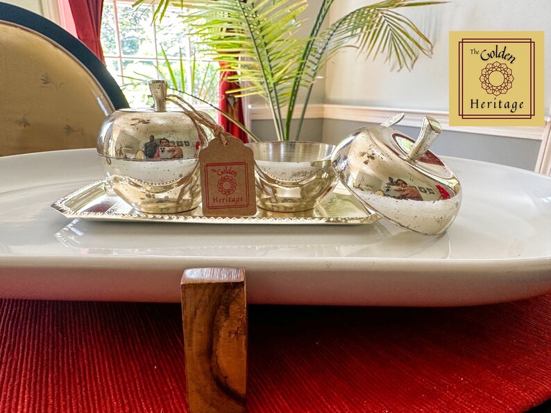 Puede incluir: Un plato de servir de plata con forma de manzana con tapa, sentado en una bandeja de plata con un borde de cuentas. El plato est&aacute; sobre una bandeja ovalada blanca, que est&aacute; sobre un mantel individual rojo. El texto "The Golden Heritage" es visible en la esquina superior derecha de la imagen.