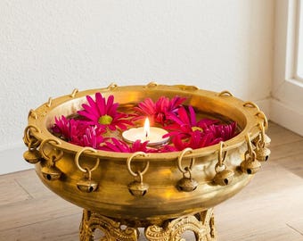 May include: A round brass bowl with a polished finish, featuring a series of small hanging bells around the rim and an ornate, decorative tripod base. The bowl is filled with water, floating magenta daisy-like flowers, and a lit tea light candle.