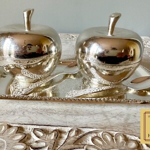 Puede incluir: Dos cuencos de plata en forma de manzana con tapas, presentados en una bandeja de plata con dise&ntilde;os ornamentados. La bandeja tiene un borde de cuentas. Los cuencos y la bandeja est&aacute;n sobre una superficie de madera con un dise&ntilde;o floral tallado.
