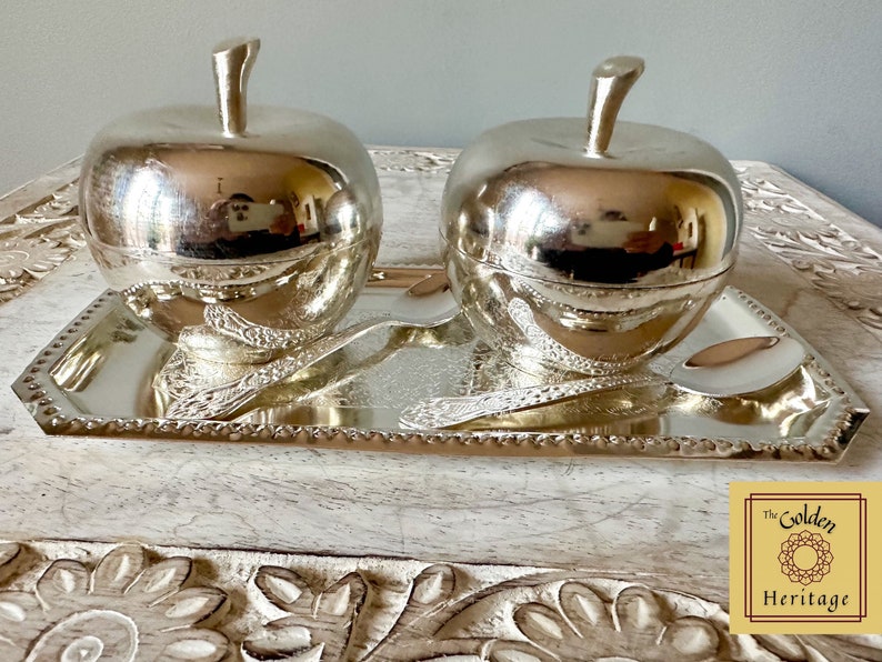 Puede incluir: Dos cuencos de plata en forma de manzana con tapas, presentados en una bandeja de plata con dise&ntilde;os ornamentados. La bandeja tiene un borde de cuentas. Los cuencos y la bandeja est&aacute;n sobre una superficie de madera con un dise&ntilde;o floral tallado.