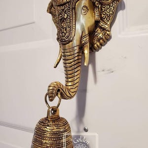 Puede incluir: Una decoraci&oacute;n de pared de cabeza de elefante de metal dorado con detalles intrincados y una campana colgante. La trompa del elefante est&aacute; enroscada y sostiene la campana. La campana tambi&eacute;n est&aacute; decorada con detalles intrincados.