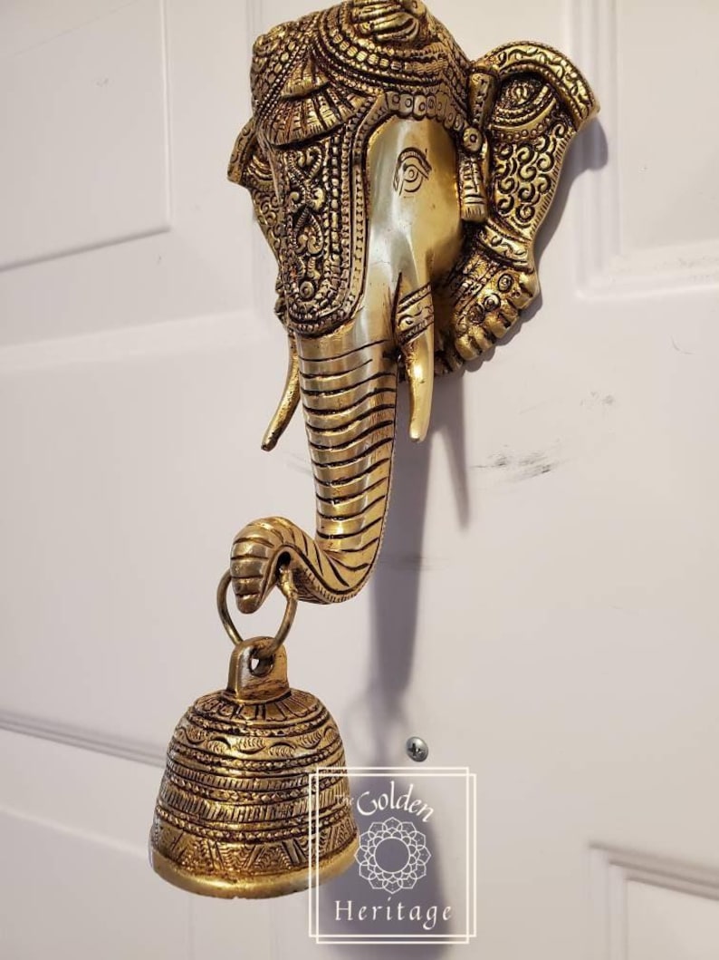 Puede incluir: Una decoraci&oacute;n de pared de cabeza de elefante de metal dorado con detalles intrincados y una campana colgante. La trompa del elefante est&aacute; enroscada y sostiene la campana. La campana tambi&eacute;n est&aacute; decorada con detalles intrincados.