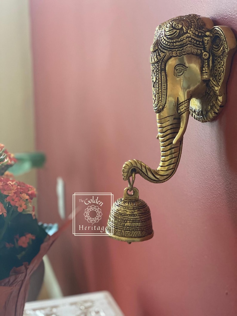Puede incluir: Una decoraci&oacute;n de pared de metal dorado con la cabeza de un elefante con detalles intrincados y una campana colgando de su trompa. La cabeza del elefante est&aacute; adornada con patrones decorativos y la campana tiene un dise&ntilde;o similar.