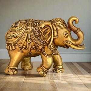 Può includere: Una statuetta di elefante dorata con dettagli intricati e la proboscide alzata. L'elefante è in piedi su una superficie di legno.