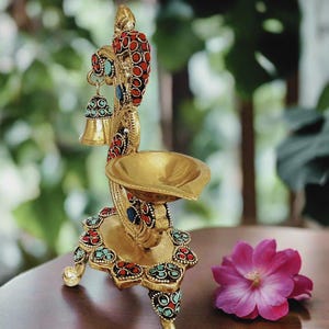 May include: A gold metal diya with intricate designs and red and blue gemstones. The diya has three legs and a bell hanging from the top.