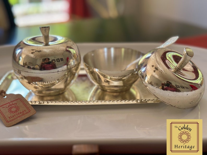 Puede incluir: Azucarero de plata en forma de manzana con tapa y un cuenco de plata a juego en una bandeja de plata. La bandeja tiene un borde de cuentas. El conjunto est&aacute; etiquetado como "The Golden Heritage".