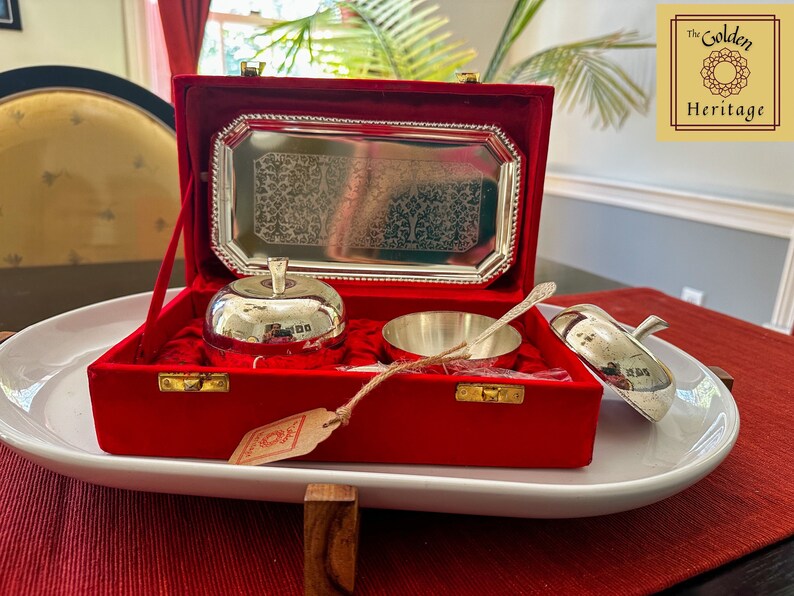 Puede incluir: Un plato de plata con forma de manzana con tapa, un cuenco de plata y una cuchara de plata se muestran en una caja de terciopelo rojo con cierre dorado. La caja est&aacute; abierta y los art&iacute;culos son visibles. La caja est&aacute; sentada sobre una bandeja ovalada blanca. El texto "The Golden Heritage" es visible en una etiqueta dorada.