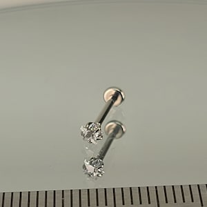 May include: Two silver-colored lip piercing studs. Each stud features a small, clear, star-shaped gemstone at the end of a straight post. The studs are displayed on a reflective surface.