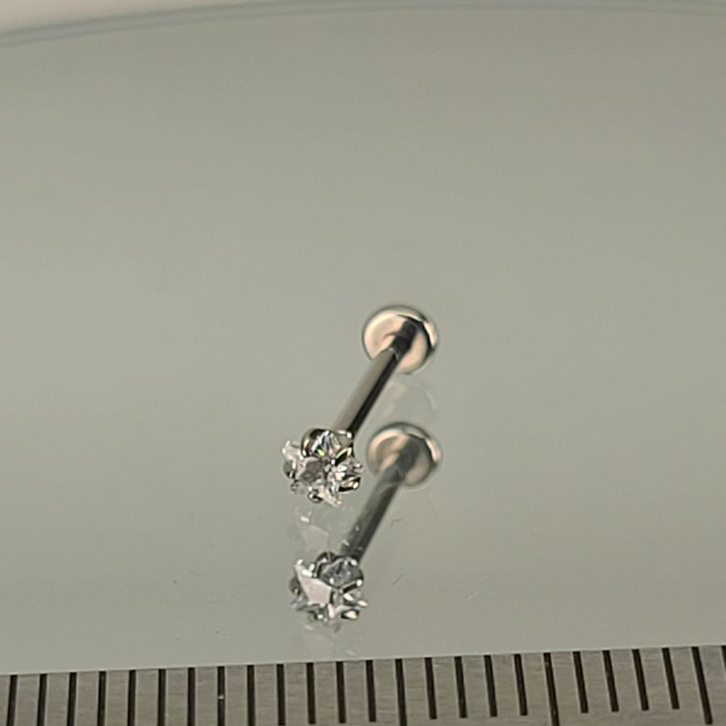 May include: Two silver-colored lip piercing studs. Each stud features a small, clear, star-shaped gemstone at the end of a straight post. The studs are displayed on a reflective surface.
