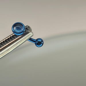 May include: Close-up of a surgical instrument holding a small, blue, spherical object. The instrument is silver and has a ridged edge. The blue object has a shiny, metallic appearance. The background is a soft, neutral color.