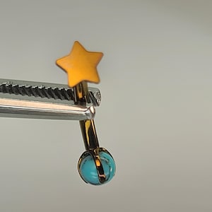 May include: A gold-colored earring with a star-shaped top and a turquoise-colored orb at the bottom. The earring is held by a silver tool, showcasing its design and details. The star is a warm yellow color.