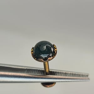 May include: A stud earring featuring a dark green, round, cabochon-cut gemstone set in a gold-toned metal prong setting. The earring has a straight post and a flat circular back, held by metal tweezers to display the front and side profile.