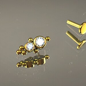 May include: A gold body jewelry piece with two clear gemstones and a row of gold beads. The jewelry is on a gray surface.