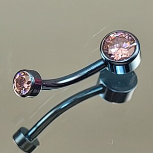 Peut inclure: Une barre de piercing corporel argentée avec deux pierres précieuses roses. La barre est incurvée et a une petite pierre précieuse à une extrémité et une plus grosse pierre précieuse à l'autre extrémité.
