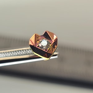 May include: A heart-shaped stud earring with a geometric design. The earring has a central, round, clear gemstone set in a rose gold and black metal frame. It is held by a silver tool.