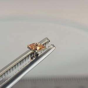May include: A marquise-cut, peach-colored gemstone held by a silver tool. The gemstone is set in a gold-colored bezel. The tool has a serrated edge and is positioned against a light background.
