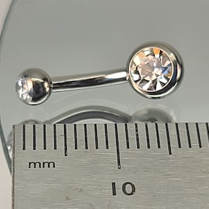 May include: Silver belly button ring with a clear crystal on each end. The ring is measured against a ruler, showing it is 10 mm long.