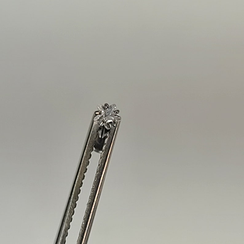 May include: A small, clear, faceted gemstone held by a silver metal tool. The gemstone has a starburst design. The tool has a serrated edge. The background is a plain, light gray.