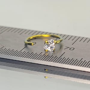 May include: A gold-colored nose ring with a clear, heart-shaped gemstone. The ring is open-ended and sits on a metal ruler, showing the measurement in millimeters. The gemstone is the focal point, reflecting light.