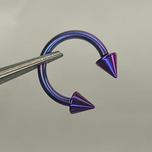 May include: A circular barbell body jewelry piece featuring an iridescent purple and blue metallic finish. The curved bar is capped with two sharp, conical spikes at each end, creating a bold, symmetrical look for various piercings.