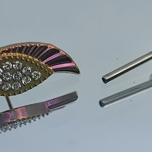 May include: A gold and pink body jewelry piece with a teardrop shape and a row of clear rhinestones. The piece comes with a silver post and two silver balls.