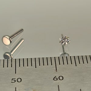 May include: Two silver-colored lip piercing studs. One stud has a flat, round top. The other stud has a starburst design with multiple facets. The studs are displayed on a reflective surface next to a ruler.