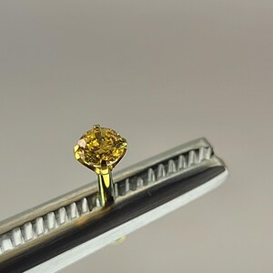 May include: A close-up of a yellow diamond ring held by a silver and gold tool. The diamond is round-cut and set in a gold prong setting. The tool has a silver body with a series of small teeth.