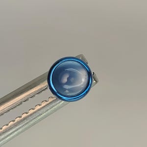 May include: A round, blue gemstone with a polished surface, set in a dark blue metal bezel. The gem is held by silver tweezers. The gemstone has a light blue hue and is likely a cabochon. The gem is approximately 1 cm in diameter.