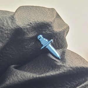 May include: A blue metal dagger pin, with a silver finish, is attached to a black fabric surface.