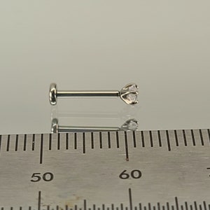 May include: A silver-colored labret stud with a clear, round gemstone. The stud has a flat back and a three-prong setting for the gemstone. The image includes a ruler for size reference.