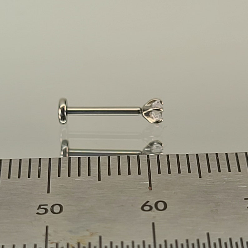 May include: A silver-colored labret stud with a clear, round gemstone. The stud has a flat back and a three-prong setting for the gemstone. The image includes a ruler for size reference.