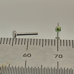 May include: Two silver nose rings with a green gemstone. The rings are shown next to a ruler with markings for 60 and 70.