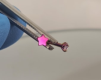 May include: A pink star-shaped opal gemstone and a clear gemstone on a surgical steel barbell, held by tweezers.