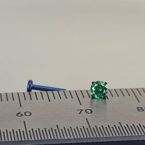 May include: A close-up of a small, green gemstone stud earring with a blue post. The earring is next to a blue post. The earring is on a ruler with measurements in millimeters.