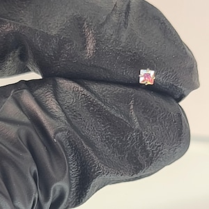 May include: A small, square-shaped earring with a pink and gold iridescent center stone. The earring is held against a black background, showcasing its delicate design and color variations. The earring is a piece of jewelry.