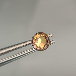 May include: A round, golden-yellow gemstone held by a pair of silver tweezers. The gemstone has a faceted surface that reflects light. The setting appears to be a rose gold color. The tweezers are in focus, and the background is a soft gray.