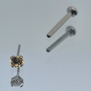 May include: A close-up of a nose piercing with a clear gem. The gem is a light yellow colour and has a round shape. The piercing is made of stainless steel.