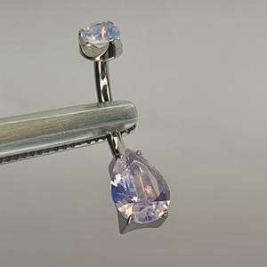 May include: Two pieces of jewellery are held by a silver tool. One piece is a small, round, light-coloured gem. The other is a larger, pear-shaped, light purple gem. Both are set in silver.