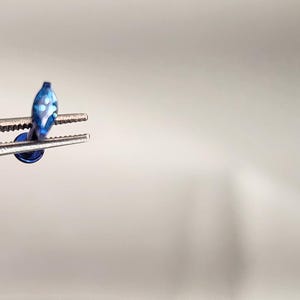 May include: A small blue body jewelry piece with a marquise-shaped top featuring white accents, held by metal tweezers. The jewelry has a flat circular base and a metallic blue finish, showcasing a slender post connecting the two components.