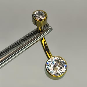 May include: A gold-colored belly button ring with two clear, round, faceted gemstones. The top gem is smaller than the bottom gem. The ring is held by a silver-colored tool.