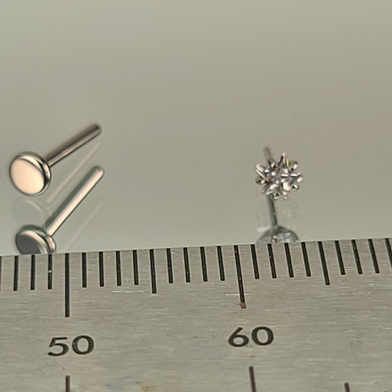 May include: Two silver-colored lip piercing studs. One stud has a flat, round top. The other stud has a starburst design with multiple facets. The studs are displayed on a reflective surface next to a ruler.