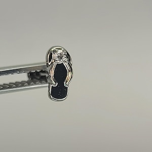 May include: A silver-toned flip-flop charm with a black sole and a clear stone accent. The charm is held by a silver tool against a neutral background. The charm is a miniature representation of a sandal.