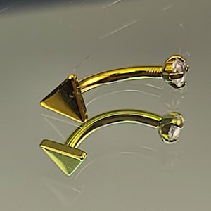 May include: Two gold-colored body jewelry pieces with a triangle-shaped end and a clear gem on the other end.