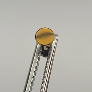 May include: A gold-colored metal button with a smooth, round surface. The button is held in place by a pair of pliers.