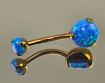 May include: A curved barbell body piercing jewelry with two blue opal-like spheres. The spheres are set in gold-colored metal, and the curved bar is also gold-colored. The jewelry is shown against a reflective surface.
