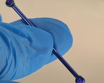 May include: A dark blue industrial barbell earring with a small, round, purple gem at each end. The jewelry is held by a person wearing a blue glove. The earring is a straight bar, designed for a piercing.