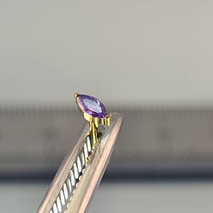 May include: A marquise-cut purple gemstone set in a gold metal setting. The gemstone is held by a pair of silver tweezers. The setting has a delicate, ornate design. The background is a light gray color.